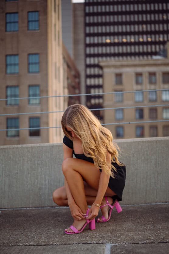 A woman with long blonde hair, wearing a black dress and pink high-heeled sandals, crouches down to adjust her shoe on a rooftop with tall city buildings in the background. by Justin Salem Meyer