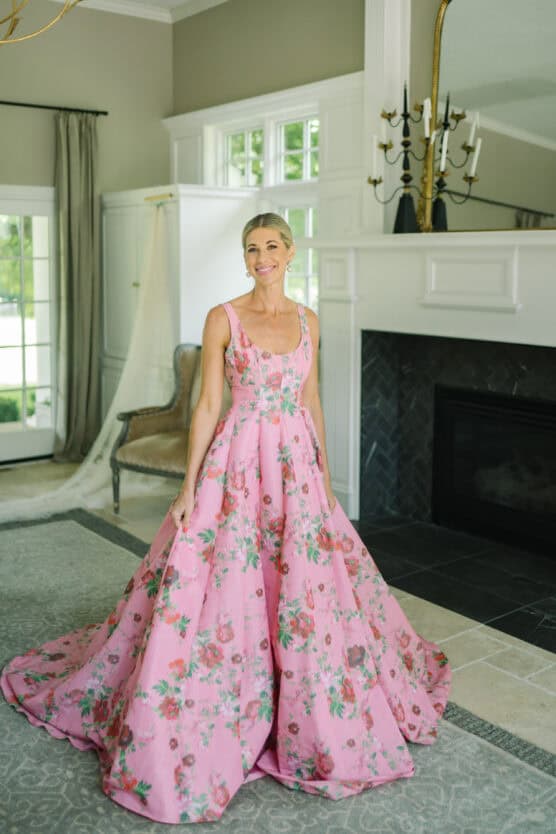 A woman stands indoors wearing a long, pink floral gown with a full skirt. She is smiling and standing on a gray rug in a bright room with large windows, a fireplace, and elegant decor. by Justin Salem Meyer