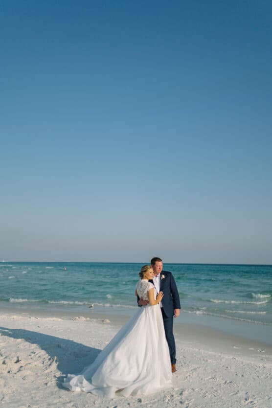 A bride and groom stand on a sandy beach near the ocean, embracing each other under a clear blue sky. The bride wears a white dress, and the groom is in a navy suit. by Justin Salem Meyer
