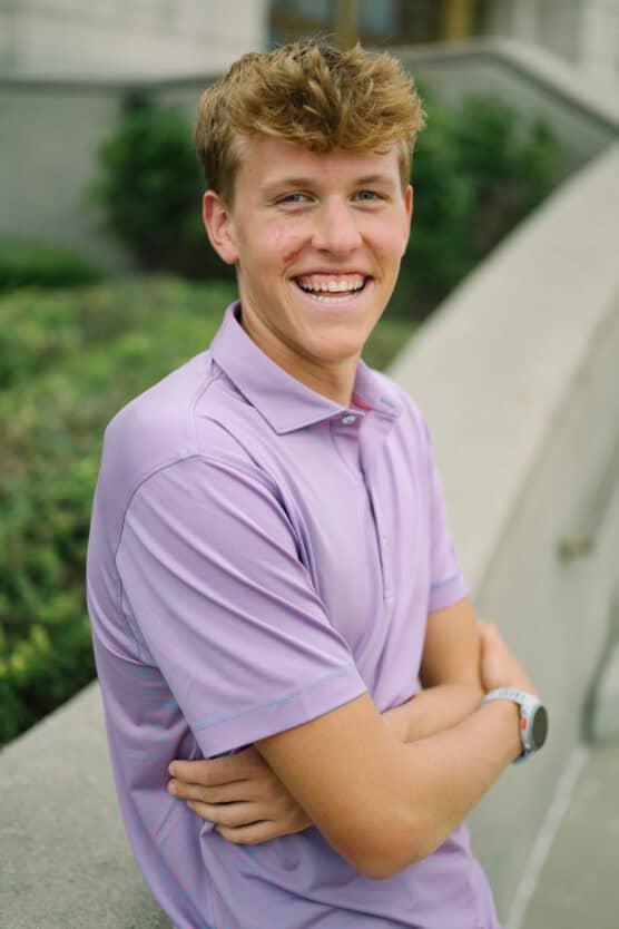 A young man with short, light brown hair smiles while sitting outdoors. He wears a light purple collared shirt and has his arms crossed, with greenery and a concrete ledge in the background. by Justin Salem Meyer