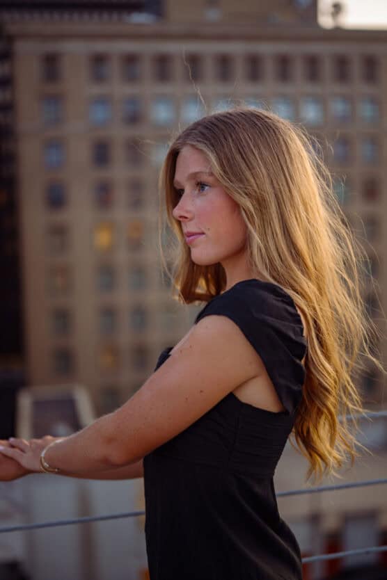 A woman with long, wavy blonde hair wearing a black dress stands outdoors, leaning on a railing, with a blurred city building in the background at sunset. by Justin Salem Meyer