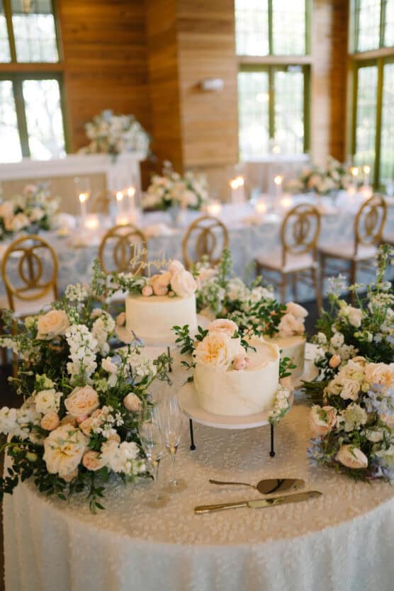 A round table decorated with white and blush floral arrangements, two white cakes topped with roses, candles, and elegant chairs in a sunlit, wood-paneled room set for a wedding reception. by Justin Salem Meyer