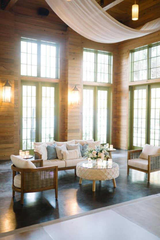 A bright, airy lounge area with wooden walls, large windows, white drapes overhead, modern wicker chairs, a white sofa, a round ottoman with flowers, and soft candlelight accents. by Justin Salem Meyer
