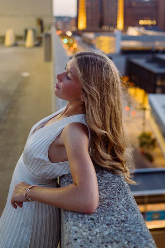 A woman with long blonde hair in a white sleeveless dress leans against a concrete ledge on a rooftop at sunset, with blurred city lights and buildings in the background. by Justin Salem Meyer