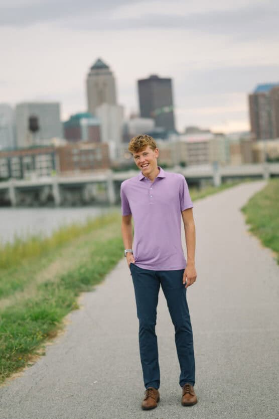 A young man in a light purple polo shirt and dark pants stands smiling on a paved path, with a city skyline and river in the background under a cloudy sky. by Justin Salem Meyer