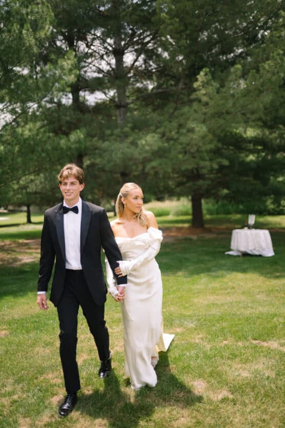 A couple dressed formally, with the man in a black tuxedo and the woman in an off-shoulder white dress, walk hand in hand on grass with tall green trees in the background. by Justin Salem Meyer
