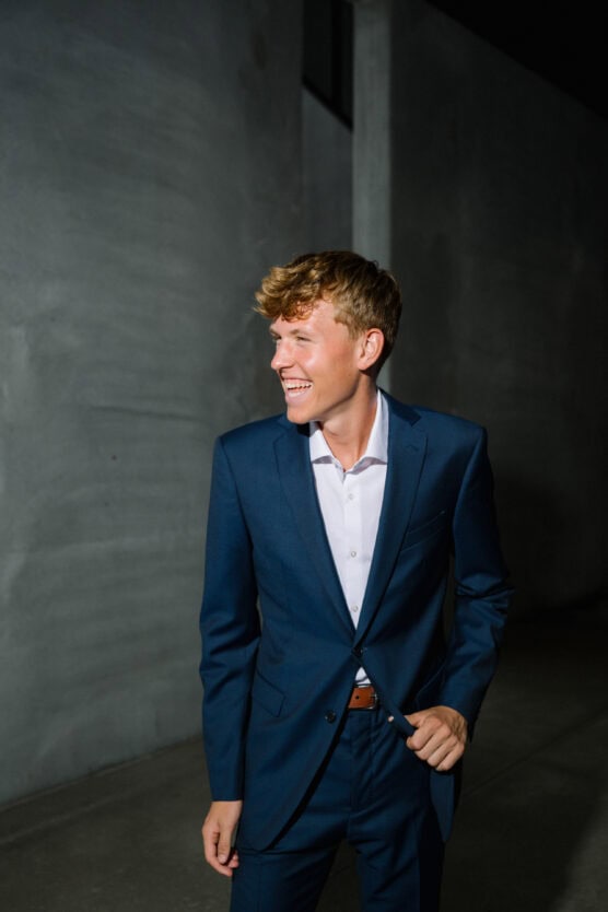 A young man in a blue suit and white shirt stands in a dimly lit, modern concrete hallway, smiling and looking to the side. by Justin Salem Meyer