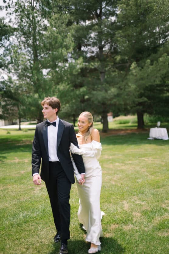 A bride in an off-shoulder white dress walks hand in hand with a groom in a black tuxedo across a grassy lawn, surrounded by tall green trees on a sunny day. by Justin Salem Meyer