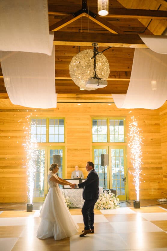 A bride and groom share their first dance in a wooden hall, with a DJ in the background and indoor fireworks sparking on either side. The room is warmly lit with large windows and draped fabric overhead. by Justin Salem Meyer