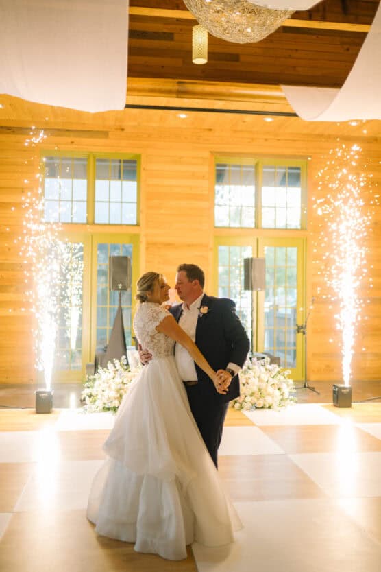 A bride and groom share their first dance in a wooden hall, surrounded by white floral arrangements and indoor fireworks, with bright windows and sparkling lights in the background. by Justin Salem Meyer