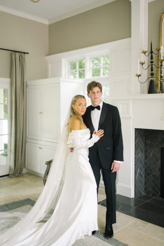 A bride in an off-the-shoulder white gown and long veil stands beside a groom in a black tuxedo. They pose indoors in an elegant room with white paneled walls, large windows, and a fireplace. by Justin Salem Meyer