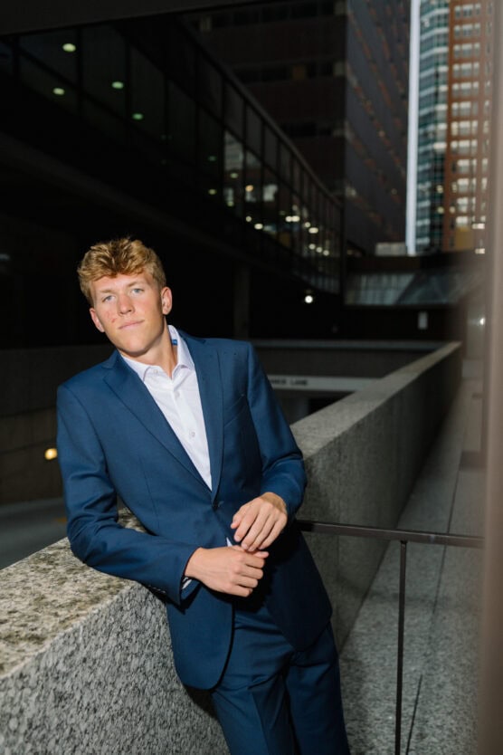 A young man in a blue suit leans against a stone railing in an urban setting at dusk, with modern buildings and lights visible in the background. by Justin Salem Meyer