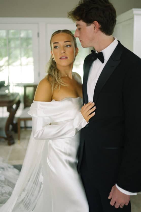A bride in an off-the-shoulder white gown stands closely with a groom in a black tuxedo. She looks away, while he gazes at her. They are indoors with natural light coming through a window behind them. by Justin Salem Meyer