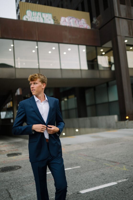 A young man in a blue suit buttons his jacket while standing on a city street. He looks to the side, with modern glass buildings and graffiti on a shipping container in the background. by Justin Salem Meyer