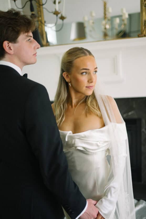 A bride in a white satin gown with a veil stands beside a groom in a black tuxedo. They hold hands and look in different directions in a bright, elegant room with a white mantel and gold mirrors. by Justin Salem Meyer