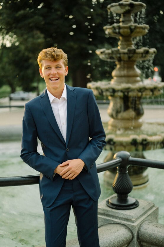 A young man in a blue suit and white shirt stands smiling in front of a stone fountain with water cascading in the background, outdoors in daylight. by Justin Salem Meyer