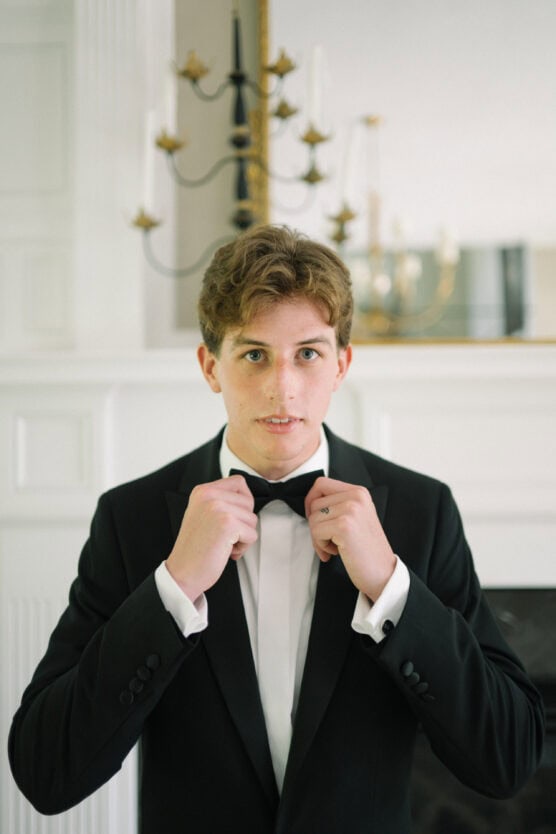A young man in a black tuxedo adjusts his bow tie while standing indoors. A chandelier and a mirror are visible in the background, giving the scene an elegant, formal atmosphere. by Justin Salem Meyer