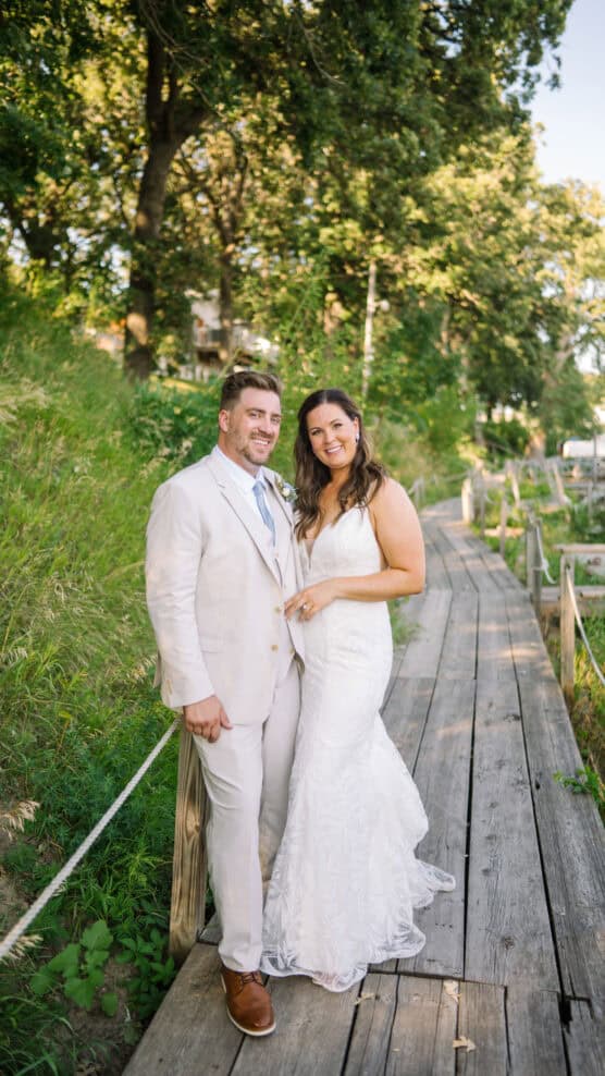 A bride and groom stand smiling on a wooden boardwalk surrounded by greenery and trees. The bride wears a white dress, and the groom wears a light suit with a blue tie. by Justin Salem Meyer