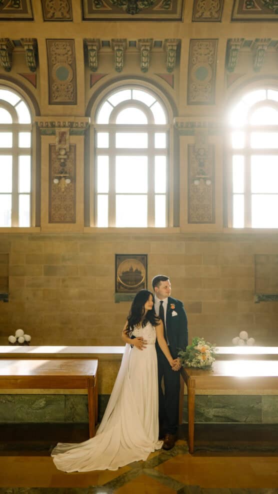A bride and groom stand together in a sunlit, elegant hall with large arched windows and ornate decorations, the bride holding her dress and bouquet while the groom holds her waist. by Justin Salem Meyer