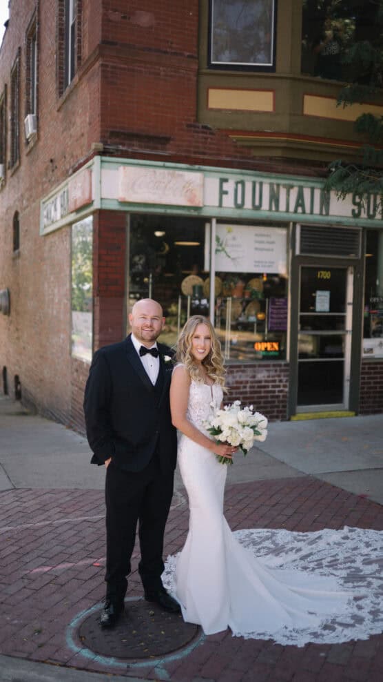 A bride in a white gown holds a bouquet and stands next to a groom in a black tuxedo outside a vintage storefront with “Fountain” on the sign. They are both smiling and standing on a brick sidewalk. by Justin Salem Meyer