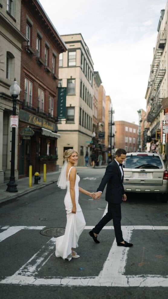 A bride in a white dress and veil holds hands with a groom in a dark suit as they cross a city street, smiling and looking back, surrounded by brick buildings and parked cars. by Justin Salem Meyer