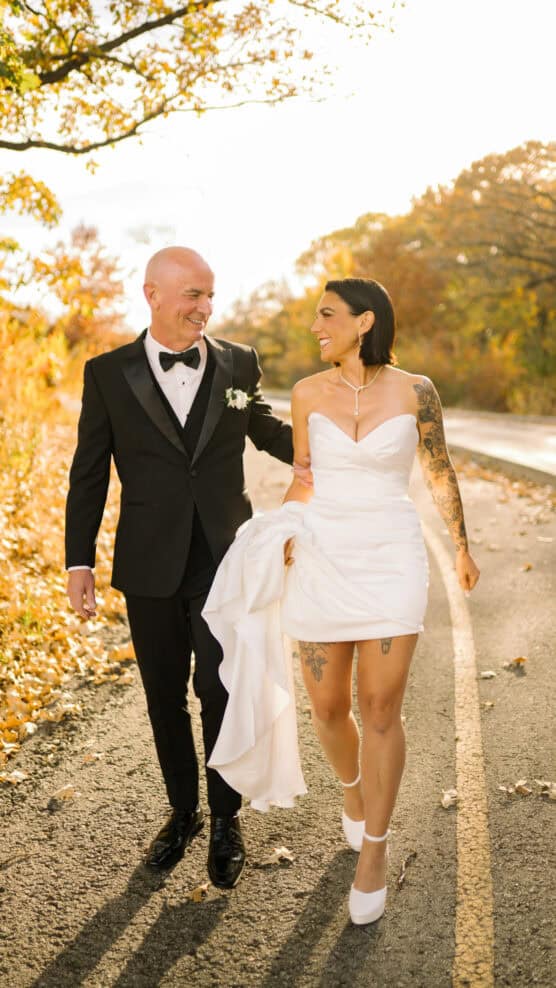 A smiling couple walks on a sunlit road surrounded by autumn trees. The man wears a black tuxedo, and the woman wears a white strapless wedding dress, holding up her skirt and looking at him. by Justin Salem Meyer