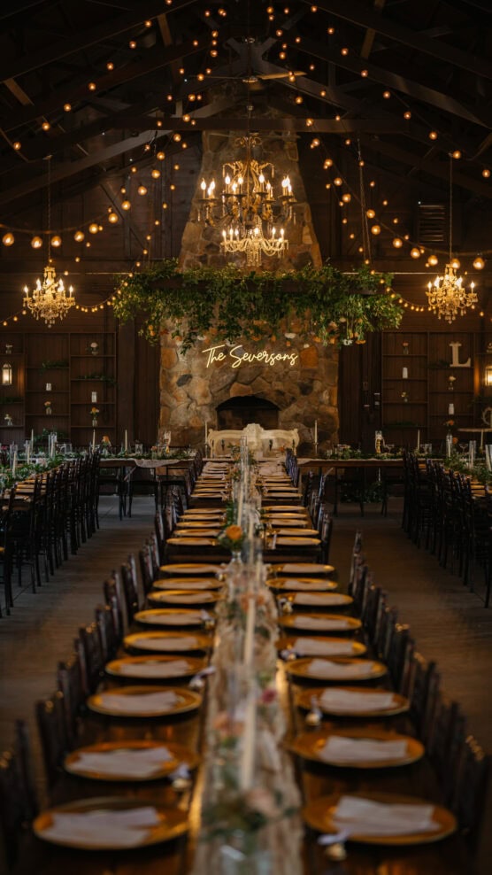 A long banquet table set for a formal event stretches toward a stone fireplace with a neon sign reading The Swensons. Chandeliers and string lights hang from the wooden ceiling, creating a warm, elegant atmosphere. by Justin Salem Meyer