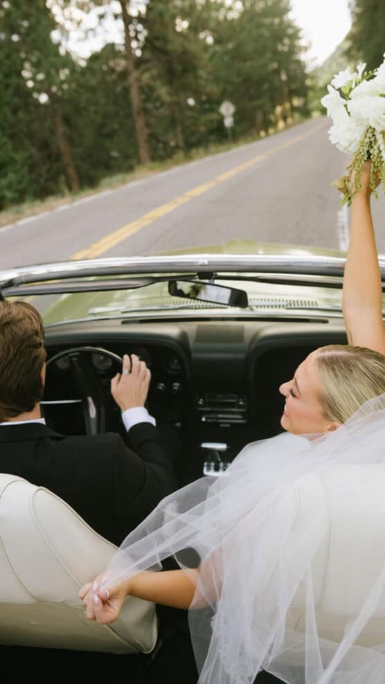 A bride and groom drive in a convertible on a tree-lined road. The bride, in a veil, holds a bouquet up joyfully while holding the groom’s hand as he drives. The photo is taken from behind them. by Justin Salem Meyer
