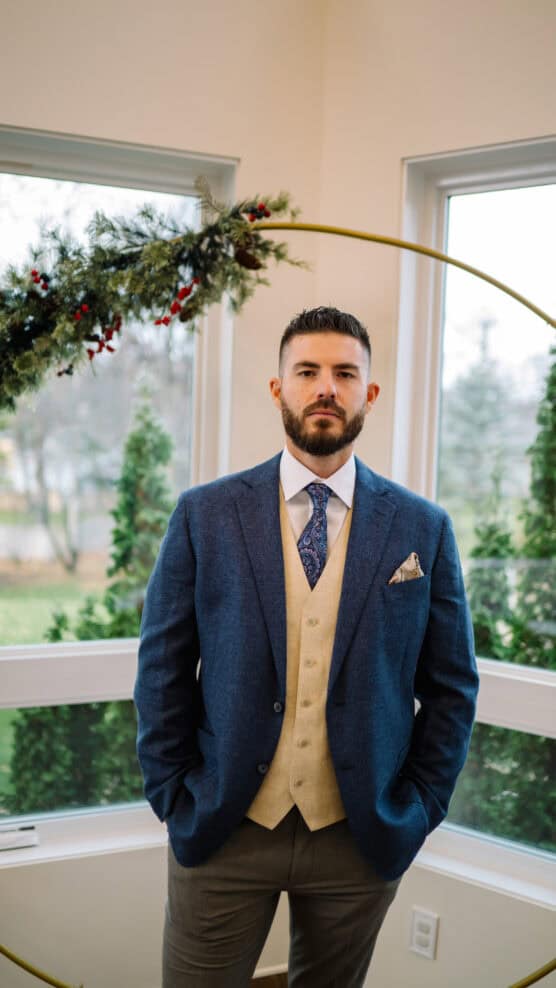 A man with a beard stands indoors wearing a blue blazer, beige vest, white shirt, patterned tie, and pocket square. Behind him are windows showing greenery and a holiday wreath with red accents. by Justin Salem Meyer