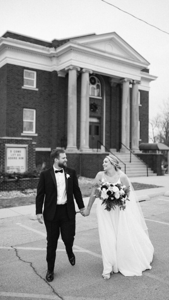 A bride and groom walk hand in hand, smiling at each other, in front of a brick building with columns. The bride holds a bouquet of flowers and wears a flowing dress, while the groom wears a black tuxedo. by Justin Salem Meyer