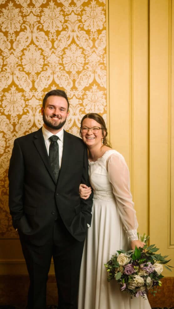 A smiling couple poses indoors; the man wears a black suit and tie, and the woman, in a white wedding dress holding a bouquet, stands beside him arm-in-arm. Ornate gold-patterned wallpaper is in the background. by Justin Salem Meyer