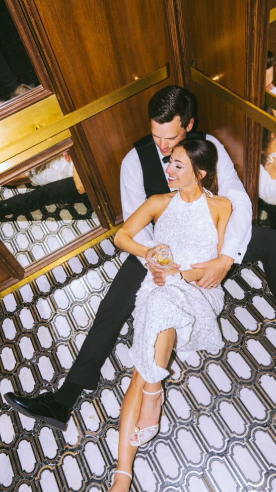 A couple in formal attire sits on the floor of an elegant elevator, smiling and embracing. The woman in a white dress holds a drink, while the man, in a vest and tie, wraps his arms around her lovingly. by Justin Salem Meyer