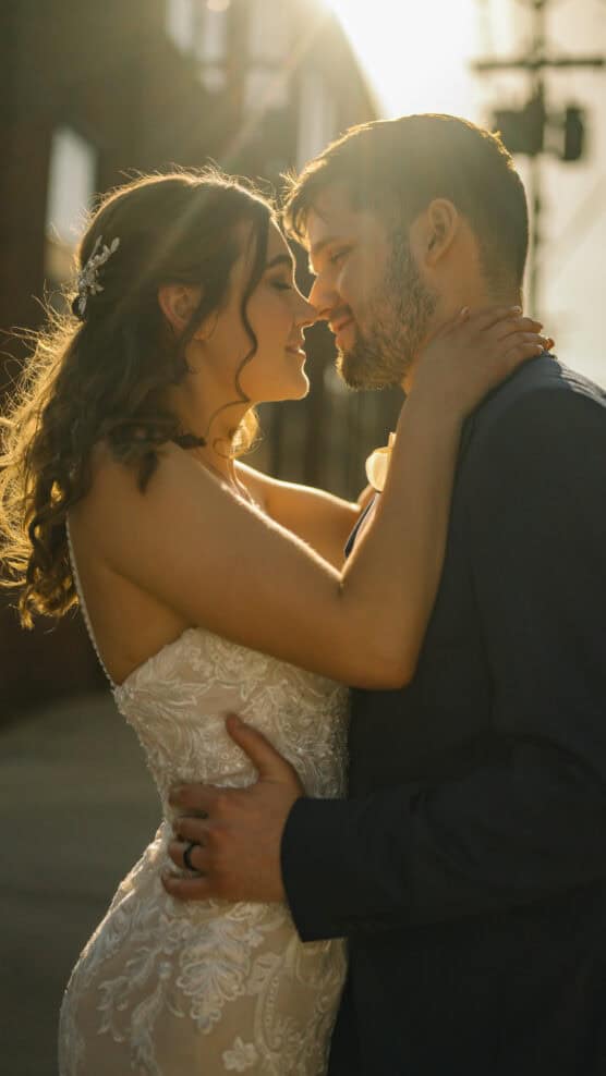 A bride and groom embrace closely outdoors at sunset, gazing into each others eyes and smiling. The bride wears a lacy white dress; the groom wears a dark suit. Sunlight creates a warm, glowing backlight around them. by Justin Salem Meyer