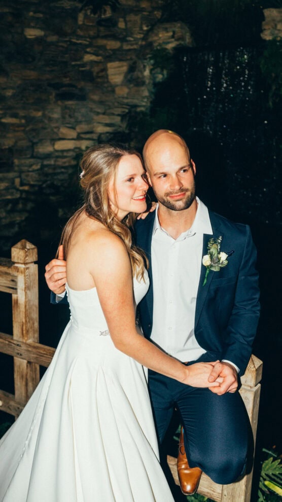 A bride in a white dress and a groom in a dark suit sit close together on a wooden fence, smiling and holding hands, with a stone wall and greenery in the background. by Justin Salem Meyer