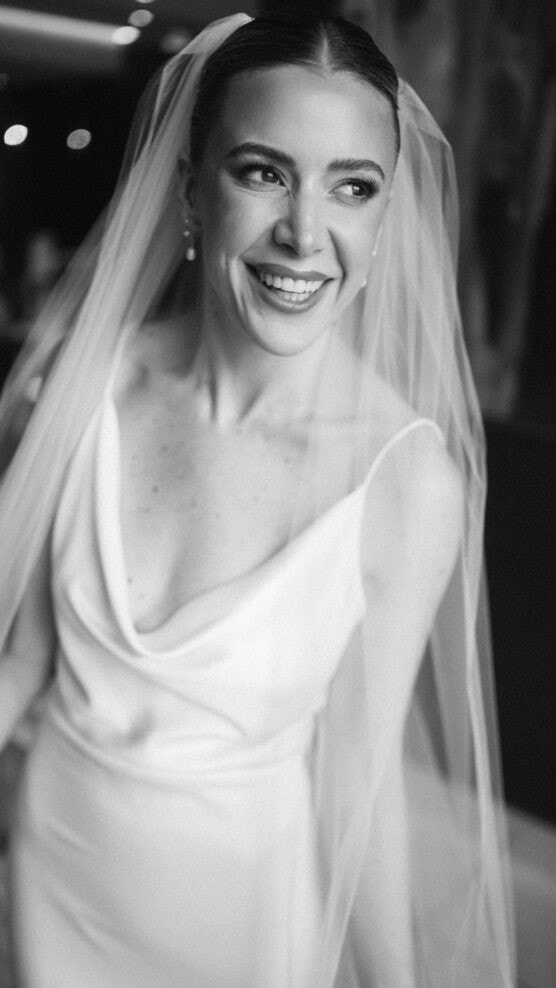 A bride in a sleeveless wedding dress and veil smiles brightly, looking away from the camera. The black and white photo captures her joyful expression and elegance. by Justin Salem Meyer