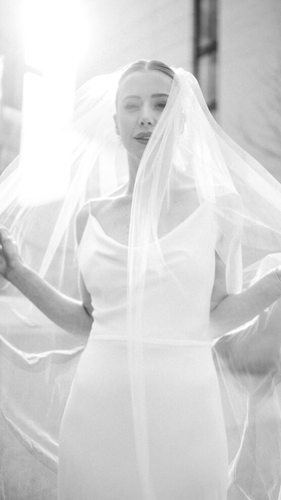 A bride in a white dress and veil poses outdoors in soft sunlight, holding her veil with both hands. The photo is black and white, creating a dreamy and romantic atmosphere. by Justin Salem Meyer