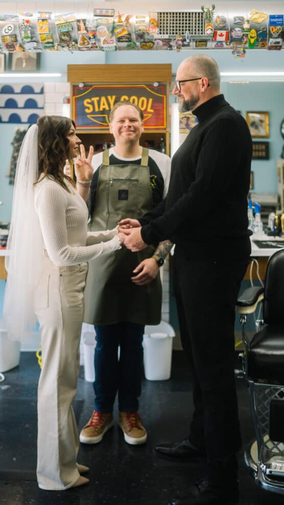 A bride and groom hold hands and smile at each other in a retro barbershop, with a smiling officiant in an apron standing behind them. The bride flashes a peace sign; colorful memorabilia line the shelf above. by Justin Salem Meyer