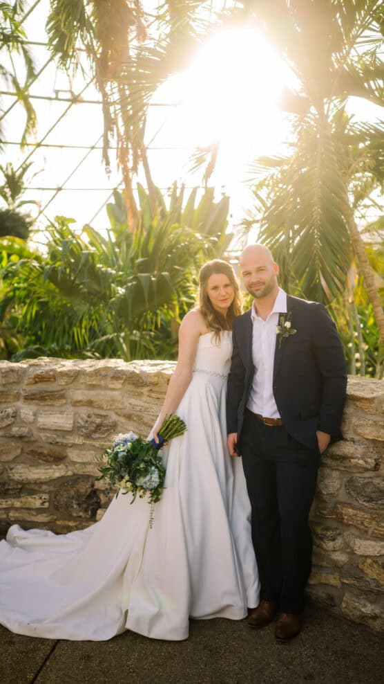 A bride in a white dress and a groom in a dark suit stand together, smiling, in front of a stone wall with lush green plants and sunlight shining through palm trees behind them. by Justin Salem Meyer