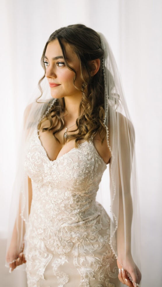 A woman in a white lace wedding dress and veil stands in soft, natural light, looking to the side with a gentle smile. Her brown hair is styled in loose curls, framing her face. by Justin Salem Meyer