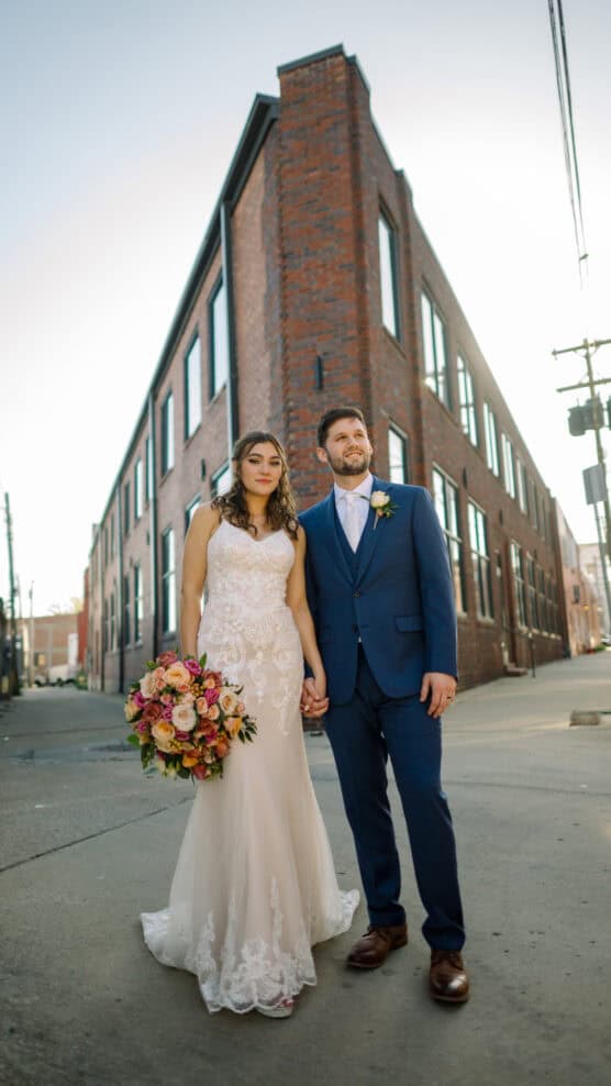 A bride in a lace wedding dress holding a colorful bouquet stands hand-in-hand with a groom in a blue suit. They are outside on a city street, with a brick building in the background. by Justin Salem Meyer