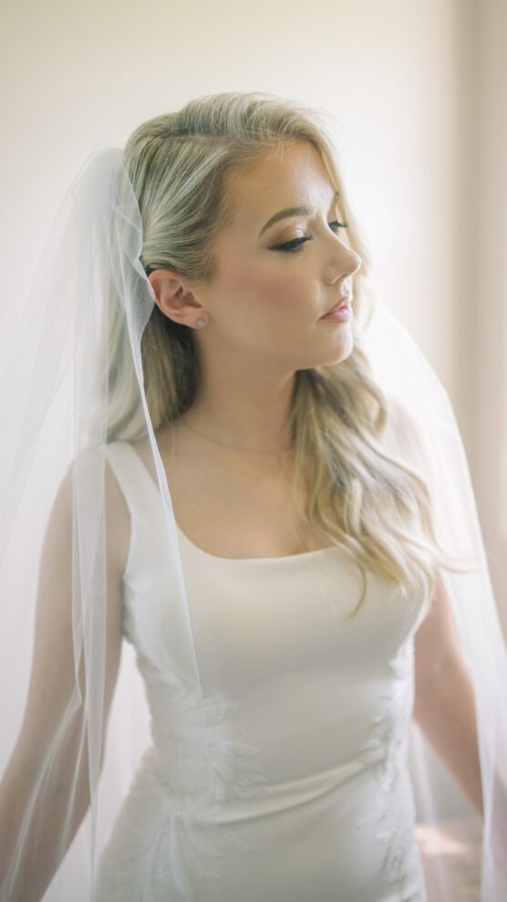 A blonde bride in a white dress and veil stands in soft natural light, looking to the side with a calm expression. by Justin Salem Meyer