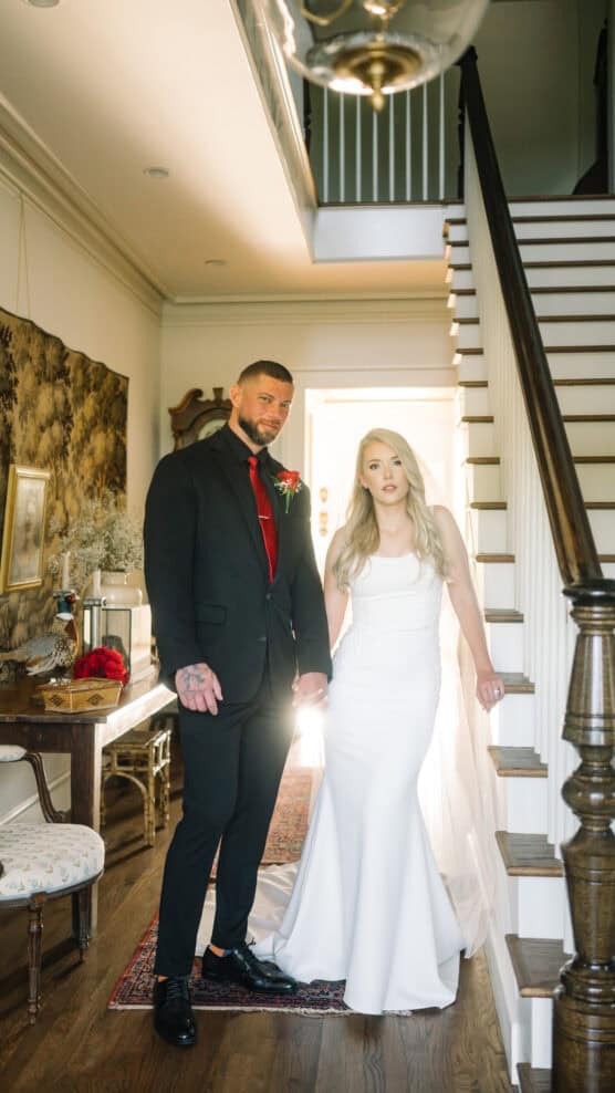 A bride in a white dress and a groom in a black suit with a red tie stand holding hands at the bottom of a staircase in a warmly lit interior with wooden floors and classic decor. by Justin Salem Meyer