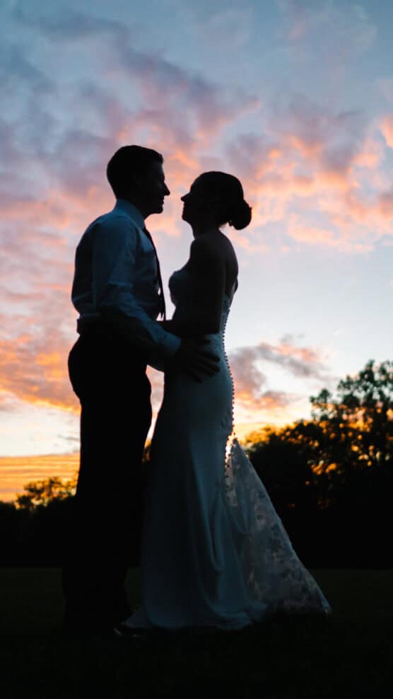 A couple in formal wedding attire stands close together outdoors at sunset, silhouetted against a colorful sky with pink, orange, and blue clouds and dark trees in the background. by Justin Salem Meyer