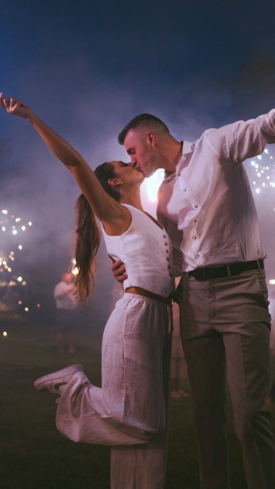 A couple dressed in white passionately kiss outdoors at night, surrounded by sparkling lights and smoke, with the woman lifting one leg in joy. by Justin Salem Meyer