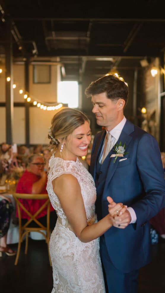 A bride in a lace dress and a groom in a blue suit share a dance and smile at a warmly lit wedding reception, with guests seated at tables in the background. by Justin Salem Meyer