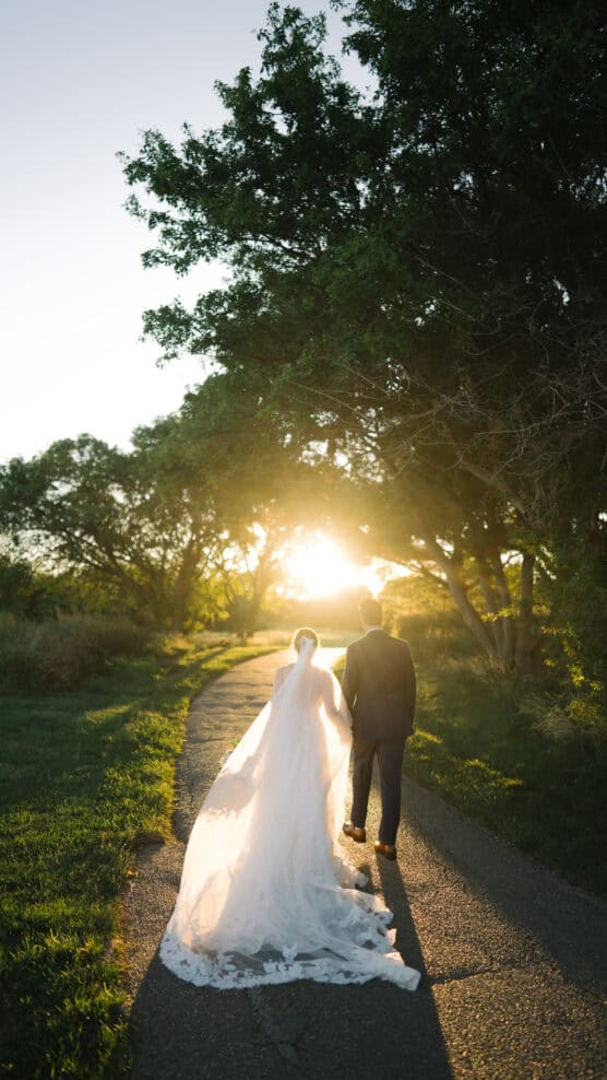 A bride and groom walk hand in hand down a sunlit path, with trees on both sides and the bride’s long veil trailing behind in the golden glow of sunset. by Justin Salem Meyer