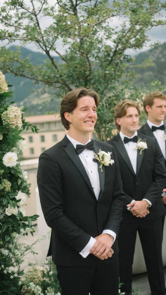 A groom in a black tuxedo with a bow tie and boutonniere stands outdoors, smiling emotionally. Two groomsmen in matching tuxedos stand behind him. There are flowers and greenery in the background. by Justin Salem Meyer