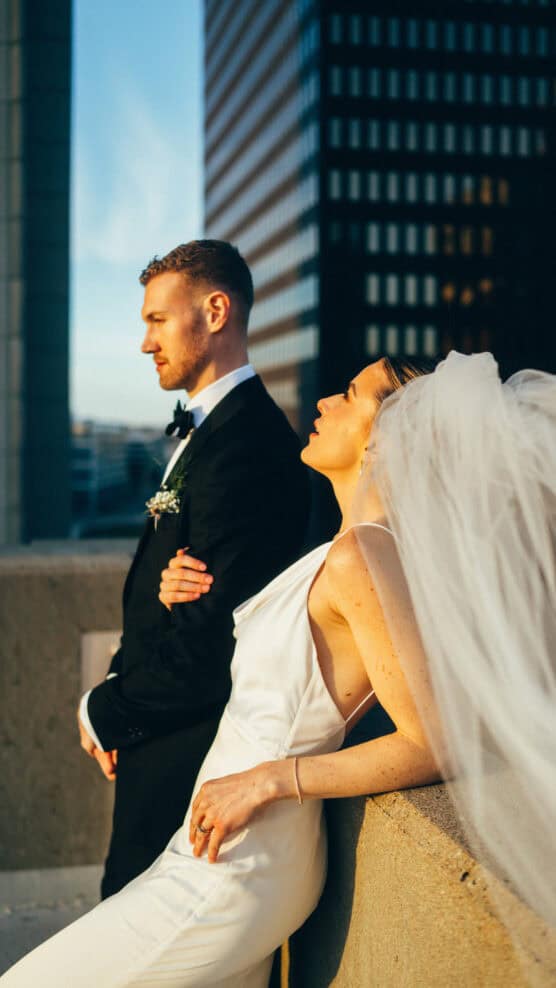 A bride in a white dress and veil leans back on a concrete ledge, eyes closed in sunlight, while a groom in a black suit stands beside her. Tall buildings and a blue sky are in the background. by Justin Salem Meyer