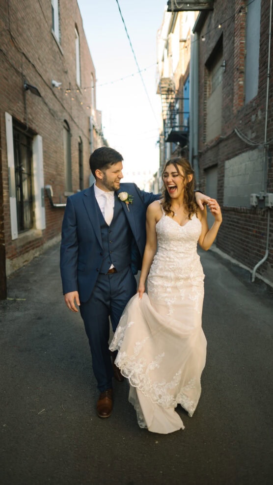 A smiling bride in a lace wedding dress walks joyfully beside her groom in a blue suit down a sunlit, brick-walled alley. They appear happy and relaxed, holding hands and laughing together. by Justin Salem Meyer